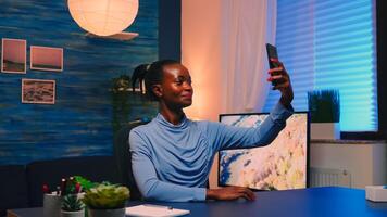 African employee taking a selfie using mobile phone smiling at camera sitting at desk in living room late at night. Black woman relaxing using modern technology network wireless doing overtime for job photo