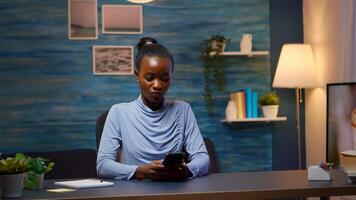 African lady taking break typing on smartphone, reading news smiling sitting at desk in living room late at night. Employee using modern technology network wireless doing overtime for job searching photo