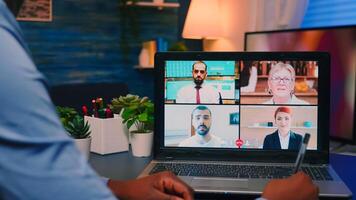 African american employee working remotely discussing with partners online using laptop sitting at desk late at night. Freelancer using modern technology network wireless talking on virtual meeting photo