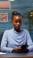 African lady taking break typing on smartphone, reading news smiling sitting at desk in living room late at night. Employee using modern technology network wireless doing overtime for job searching photo