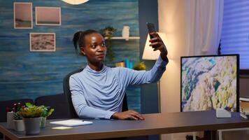 African woman taking a selfie using smartphone smiling sitting at desk in living room late at night. Black employee relaxing using modern technology network wireless, doing overtime for job photo