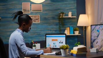 African entrepreneur checking financial project of company looking on laptop analysing annual statistics data. Taking notes sitting at desk in living room office late at night overtime. photo