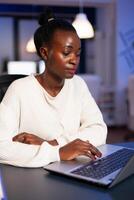 Stressed african manager woman working with financial graphs checking statistics, typing on laptop, reading raports late at night in start-up office doing overtime to respect deadline of project. photo