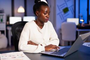 Busy african manager working on financial reports checking statistics graphs, typing on laptop sitting at desk late at night in start-up office doing overtime to respect deadline of financial project. photo