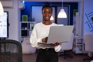 Black business woman looking at camera smiling holding laptop standing near desk in start-up company late at night. Focused employee using technology network wireless doing overtime for job. photo