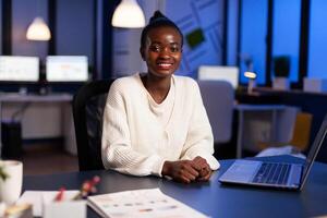 Close up portrait of business african woman smiling at camera working using laptop sitting in startup company late at night. Focused employee doing overtime respecting deadline of project. photo