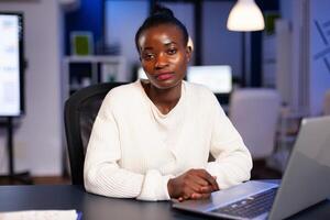 Focused tired business woman looking at camera after reading tasks on laptop sittting at desk in start-up company late at night. Employee doing overtime for job respecting deadline photo