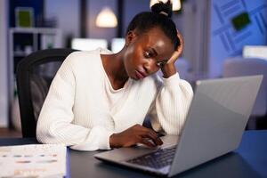 Exhausted african freelancer resting head on hand in front of laptop working overtime in start up company office. Stressed employee using modern technology doing overtime for deadline of project. photo