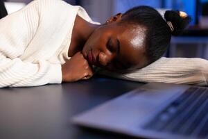Exhausted overload african business woman falling asleep on desk with open laptop monitor while working in start up company. Overworked employee using doing overtime respecting deadline. photo