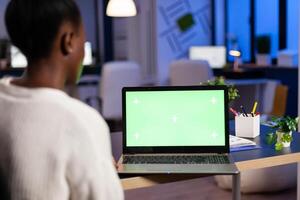 Dark skin freelancer doing overwork in office with green screen latop sitting at desk in business office late at night. Businesswoman watching desktop with green mockup, chroma key, working overtime. photo