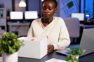 African woman being upset because she got fired from company, dismissed. Unemployed packing things late at night. woman leaving workplace office in midnight. photo