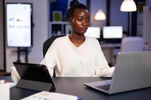 Workaholic african manager trying to finish deadline checking charts on tablet pc and laptop. Busy multitasking employee analysing financial statistics overworking writing, searching. photo