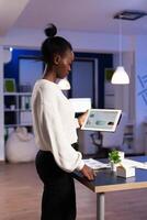 Exhausted african businesswoman trying to finish deadline working with charts on tablet pc. Busy african employee analysing financial statistics in workplace overworking writing, searching. photo