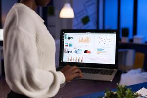 Back view of african business woman analysing financial statistics holding laptop late at night. Busy focused employee using modern technology network wireless overtime. photo