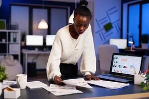 Stressed african manager woman working with financial documents standing at desk checking graphs, holding papers, reading raports late at night in start-up office doing overtime to respect deadline. photo