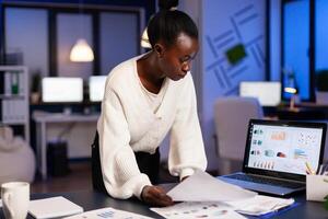 Stressed african manager woman working with financial documents checking graphs, holding papers, reading raports, standing at desk late at night in start-up office doing overtime to respect deadline. photo
