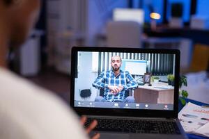African businesswoman having corporate meeting with remotely paralyzed man using webcam of laptop sitting in start up business office working overtime. Manager talking on conference at midnight. photo