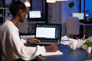 African architect working late at night on laptop with blue prints, looking at graph. Industrial woman employee studying prototype idea showing cad software on device display. photo