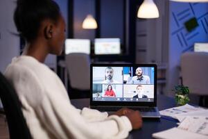 African employee working overtime from business office late at night discussing with partners online using webcam. Diverse team talking during corporate meeting using technology network wireless. photo