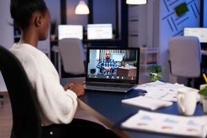 African employee working overtime from business office late at night discussing with partners online using webcam. Diverse team talking during corporate meeting using technology network wireless. photo