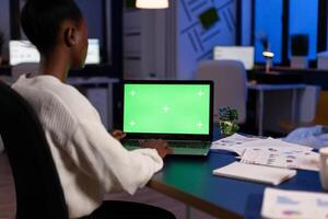 African manager typing at laptop with chroma key display during night time in start up business office working overtime. Freelancer watching mockup desktop monitor with green screen, writing data. photo