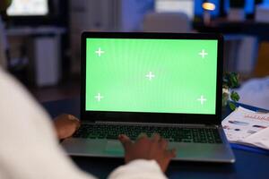 Black freelancer working on laptop with green screen display, green mockup, chroma key desktop sitting at desk in business office working at night. Freelancer watching at isolated desktop overwork. photo