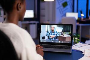 Black woman freelancer discussing with paralyzed client on call at midnight from business office using headphone. Businesswoman using virtual conference talking on webcam during online meeting. photo