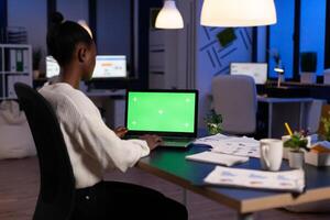 Dark skin freelancer working on laptop with green screen display sitting at desk in business office late at night. Businesswoman watching desktop with green mockup, chroma key, working overtime. photo