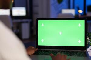 Back view of black business woman using laptop with green screen, green mockup, chroma key desktop sitting at desk in start up business office overworking. Freelancer watching at isolated display. photo