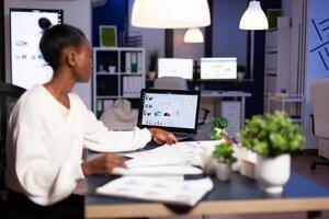 African woman working late at night looking at charst on laptop. Focused employee doing overtime for job respecting deadline of project writing, searching. photo