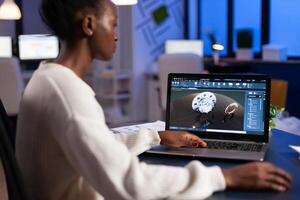 Tired african designer engineer analyzing new prototype of 3D model of industry product, working overtime. Industrial female worker studying gears idea on laptop showing cad software on device display. photo