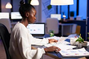 Black manager analysing graphics and financial statistics from documents working overtime. Focused employee doing overtime for job respecting deadline of project writing, searching. photo