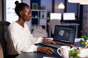 African woman engineer working at new project using laptop doing overtime at night sitting at desk in start-up office. Industrial construction designer studying on pc showing cad software in midnight. photo