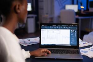 Over shoulder view of african architect working late at night in office using cad software, house blue prints. Industrial woman employee studying prototype idea showing cad software on device display. photo