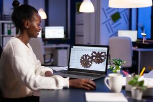 African engineer working late at night on a 3D model of industrial gears at laptop doing overtime in start up office. Freelancer studying prototype idea on pc showing cad software on device display. photo