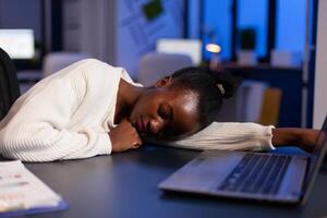 Tired exhausted african american businesswoman sleeping on desk table late at night in startup office after working at management statistics. Workaholic black manager planning company strategy photo