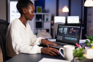 Black delighted videographer editing movie on professional laptop sitting at desk in business office at midnight. Creative editor working at night at new project processing audio film montage. photo
