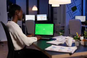 Concentrated african american businesswoman hardworking at financial strategy late at night in startup company office. Mock up green screen chroma key laptop with isolated display standing on table photo