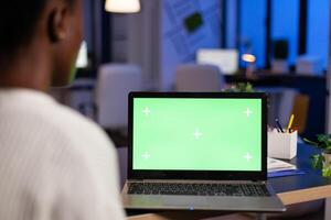 Tired african american businesswoman looking at mock up green screen chroma key laptop with isolated display overworking in startup company office late at night. Manager planning financial strategy photo
