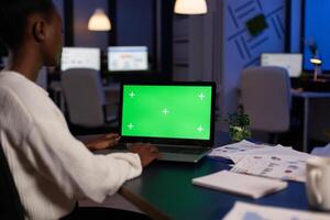 Concentrated african american businesswoman hardworking at financial strategy late at night in startup company office. Mock up green screen chroma key laptop with isolated display standing on table photo