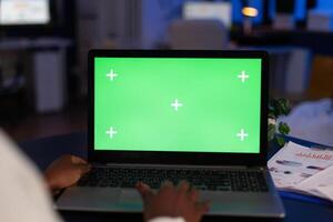 Mock up green screen chroma key laptop computer with isolated display standing on table in startup company office. African american businesswoman working at marketing project late at night photo