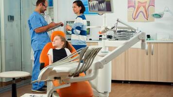 Nurse inviting kid patient in consultation dental room, pediatric doctor speaking with little girl. Man assistant talking with mother preparing for stomatologic examination in orthodontist office photo