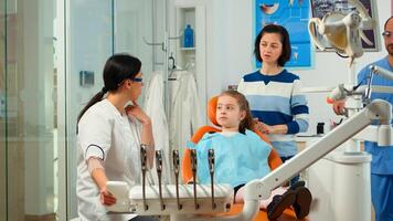 Stomatologist doctor explaining to mother the dental process of intervention for teeth problems of kid, girl showing to affected mass. Orthodontist speaking to child sitting on stomatological chair photo