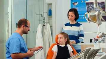 Friendly man dentist assistant interrogating kid patient and taking notes on clipboard waiting for doctor. Mother of little girl explaining dental problem to nurse indicating at toothache problems. photo