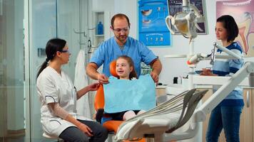 Little girl patient with tooth pain explaining dental problem to pediatric dentist and indicating at toothache with tongue. Stomatologist taking with mother about stomatologic examination of kid. photo