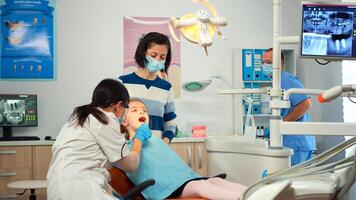 Dentist technician with gloves explaining the process to kid patient before surgery lighting the lamp. Orthodontist speaking to girl with toothache sitting on stomatological chair. photo