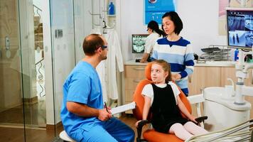 Nurse taking notes on clipboard about little girl dental problems waiting for orthodontist. Assistant explaining medical procedure to her mother standing near stomatological chair. photo