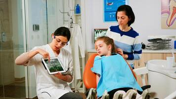 Orthodontist holding tablet and showing jaw x-ray to little patient while suggesting treatment during consultation. Stomatologist presenting to mother of girl dental radiography using modern gadget. photo