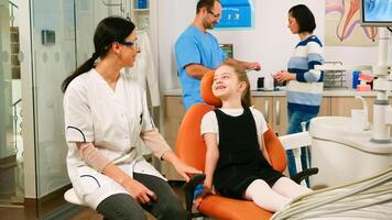 Little girl patient waiting pediatric stomatologist woman sitting in dental chair while man assistant talking with mother in background. Dentist discussing with girl before examining oral health. photo