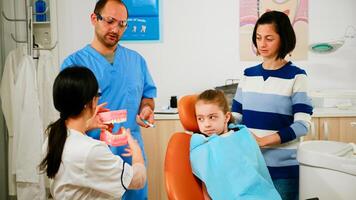 Doctor showing to child the correct dental hygiene using mock-up of teeth skeleton. Dentist holding sample of human jaw with toothbrush in stomatology office working with man assistant. photo